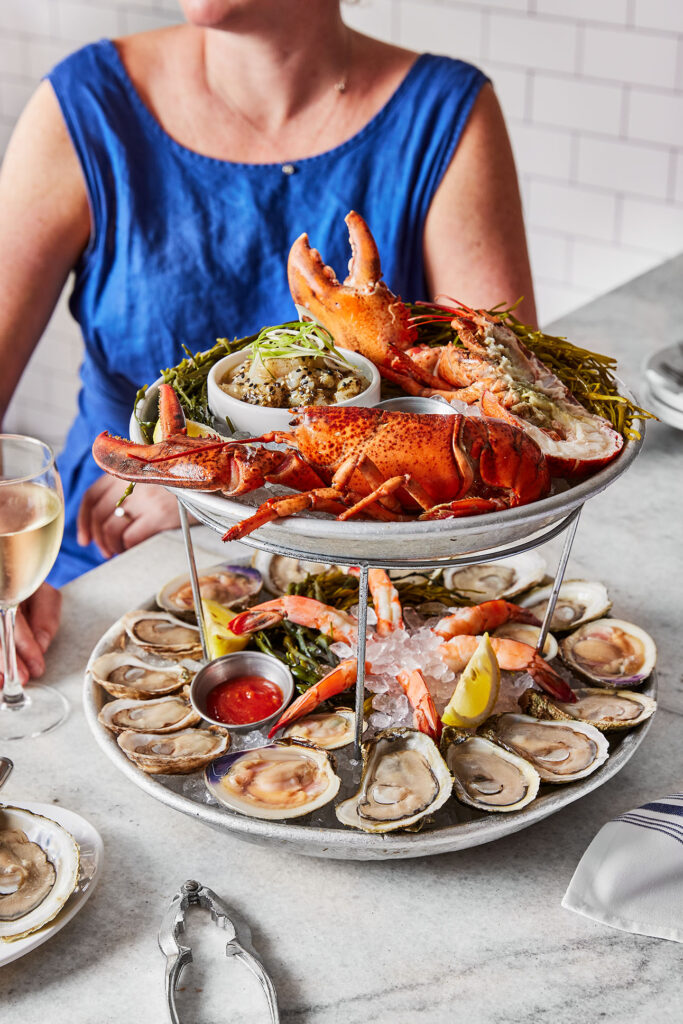 A photo of a woman sitting on a table in front of a two-tier tray full of seafood. From Lobster to shrimp to oysters served over ice.