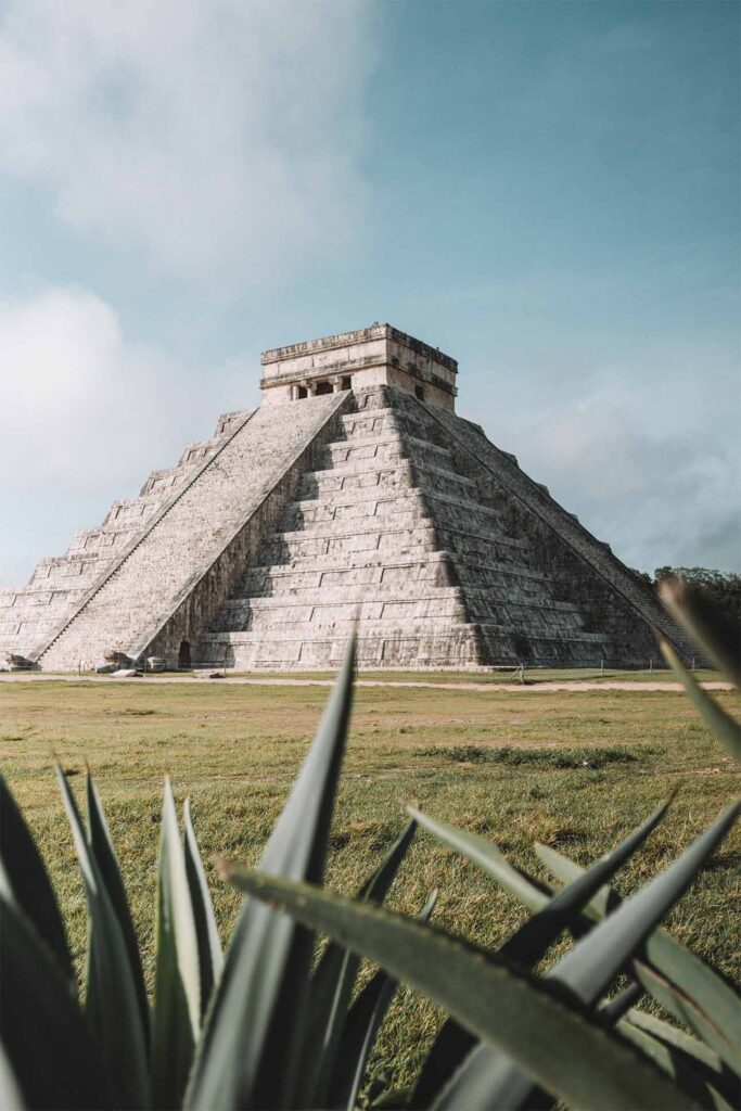 a pyramid with a stone structure in the middle of a grassy field with Chichen Itza in the background